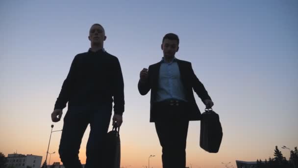 Two happy young colleagues with briefcases funny dancing while walking after successful business deal at city street. Cheerful businessmen celebrating achievement outdoor in evening. Low angle view — Stock Video