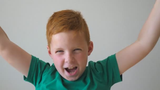 Όμορφο αγόρι κοκκινομάλλα με φακίδες κοιτώντας την κάμερα και να αυξήσει τα χέρια που χαίρεσαι μέσα επίτευξης. Πορτρέτο της ευτυχισμένη παιδί αρπάζοντας το κεφάλι του και να δείχνει χαρά στο πρόσωπό του. Αργή κίνηση — Αρχείο Βίντεο