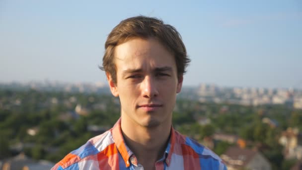 Retrato de um jovem feliz sentado no telhado de um edifício alto com uma paisagem urbana turva. Um tipo bonito a olhar para a câmara e a rir. Cara de humano alegre. Fechar câmera lenta — Vídeo de Stock