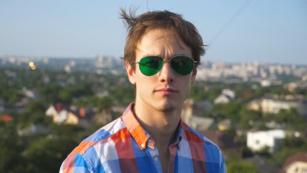 Крупный план портрета молодого человека в солнцезащитных очках, сидящего на крыше высотного здания с городским пейзажем. Красивый парень уверенно смотрит в камеру с красивым размытым пейзажем. Медленное движение — стоковое видео