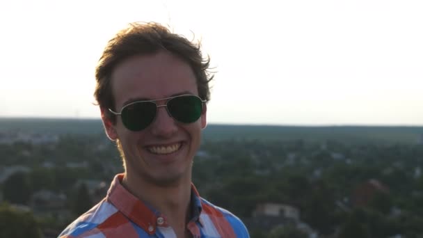 Retrato del joven hombre feliz de pie en la azotea del edificio de gran altura con un fondo borroso del paisaje urbano. Un chico guapo con gafas de sol mirando a la cámara y sonriendo. Dolly disparo Primer plano cámara lenta — Vídeos de Stock