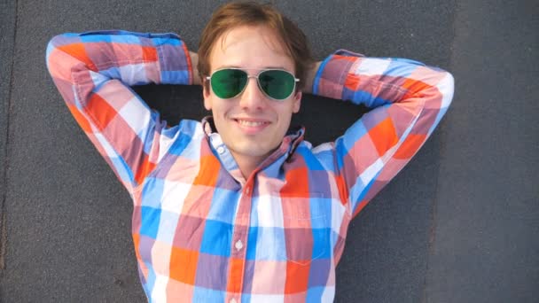 Visão superior do jovem homem feliz em óculos de sol deitado no telhado do edifício de arranha-céus e sorrindo. Bonito cara de camisa olhando para a câmera e gostando de passar tempo no telhado. Fechar câmera lenta — Vídeo de Stock