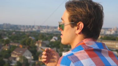 Profil sahibi çatı kenarındaki sigara ve yüz buruşturma gösteren güneş gözlüğü mutlu adam. Genç yüksek katlı bina ve rahatlatıcı çatı üzerinde duran adam. Arka plan, bulanık cityscape