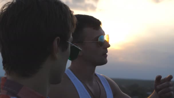 Perfil de jovem casal masculino em óculos de sol em pé na borda do telhado e falando. Bonitos meninos gays relaxando no telhado do edifício de arranha-céus e desfrutando de bela paisagem urbana. Fechar câmera lenta — Vídeo de Stock