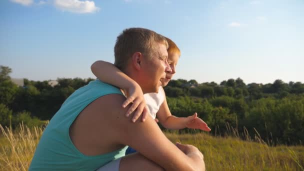 Papà e bambino seduto a erba verde sulla collina e guardando alla bella natura in avanti loro. Padre con il suo figlioletto passare del tempo insieme all'aperto al prato. Chiudere rallentatore — Video Stock