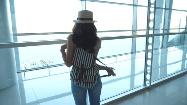 Młoda kobieta w kapeluszu plecak stojąc w terminalu lotniska i patrząc w okno. Dziewczyna czeka na jej samolot, a następnie liści. Pojęcie turystyki i podróży. Widok z tyłu powrotem zwolnionym tempie — Wideo stockowe