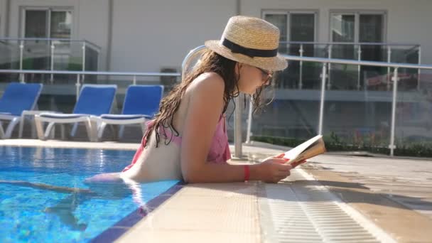 Vacker flicka i hatt och solglasögon bok vid poolen. Ung kvinna avkopplande på varm solig dag under semester. Sommar semester koncept. Sidovy närbild Slow motion — Stockvideo