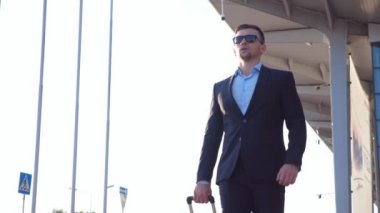 Yakışıklı genç işadamı şehir caddesindeki havaalanından bavulla geliyor. Siyah takım elbiseli kendinden emin bir adam şehir ortamında yürüyor ve tekerlekli bavulu çekiyor. Düşük açı görünümü Yavaş çekim