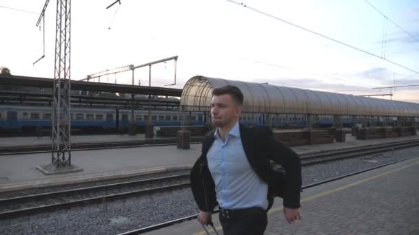 Красивый бизнесмен бежит через железнодорожную станцию спеша на поезд. Успешный бизнесмен бегает с багажом возле железной дороги. Молодой человек опаздывает на поезд. Концепция деловой поездки. Медленно — стоковое видео