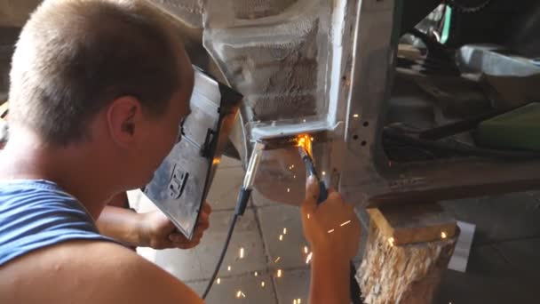 Mannelijke automaster lassen metalen delen van oude auto met lassen machine in garage. Adult reparateur of monteur werknemer doen lassen werken met behulp van professionele apparatuur en beschermende masker binnen. Dolly schot — Stockvideo