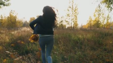 Genç bir kadın elinde sarı yapraklarla sonbahar ormanlarında koşuyor ve onları fırlatıyor. Renkli sonbahar ormanlarında eğlenen bir kadın. Gün batımında dışarıda koşan bir kız. Yavaş çekim