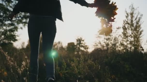 Mladá žena běží podzimním parkem s kyticí žlutých javorových listů v ruce. Dívka se baví v barevném podzimním lese s živým spadlým listím. Slunce osvětluje životní prostředí. Zpomalený pohyb — Stock video