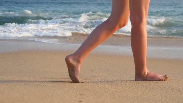 Pies femeninos caminando sobre arena dorada en la playa con olas oceánicas al fondo. Piernas de mujer joven pisando arena. Chica descalza en la orilla del mar. Vacaciones de verano. Cámara lenta Primer plano — Vídeo de stock