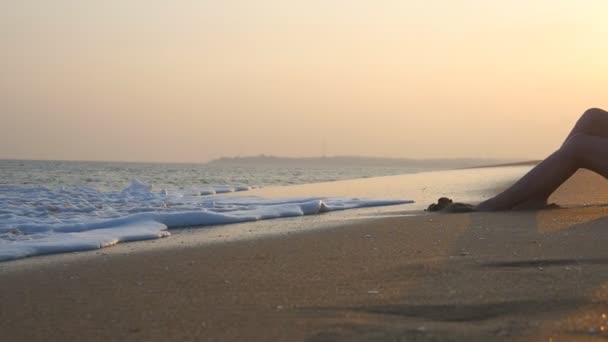 Ondas do oceano a lavar-se sobre o corpo feminino bronzeado. Menina bonito deitado na praia nas ondas na hora do pôr do sol. Mulher bonita relaxando na costa do mar durante as férias de verão. Lento movimento Fechar — Vídeo de Stock