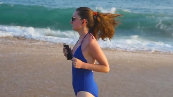 Jonge vrouw in badpak rennen op zee strand en luisteren muziek. Meisje joggen langs Ocean Shore. Vrouwelijke sportman die buiten traint. Gezonde actieve workout Lifestyle in de natuur. Slow-motion sluiten — Stockvideo