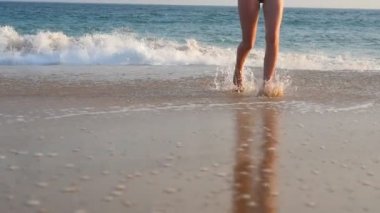 Okyanustan kum plaja yürüyen genç kadının bacakları. Bikinili tanınmayan kızın ayakları denizden kıyıya doğru adım atıyor. Yaz tatili ya da tatil. Dinlenme kavramı. Yavaş çekim