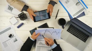 İki başarılı iş ortağının erkek elleri grafikleri inceler ve ofiste finansal raporları tartışırlar. Genç işadamları masada oturuyor ve yeni bir iş projesi geliştiriyor. Üstte görüntü