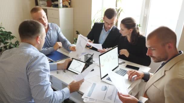 近代的なオフィスでのチームミーティング中に収入チャートやグラフを議論ビジネスの人々.若い同僚がテーブルに座って財務報告を分析しています。机の上で書類を調べる同僚 — ストック動画