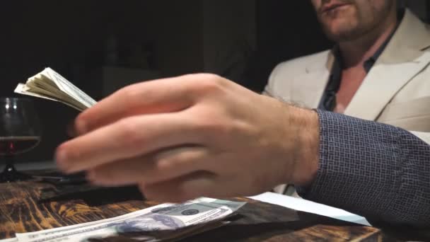Close up hands of drug dealer holding cash and counting foreign currency over table. Male arms of businessman considers one hundred dollar bills over desk and drinking brandy from glass. Slow motion — Stock Video