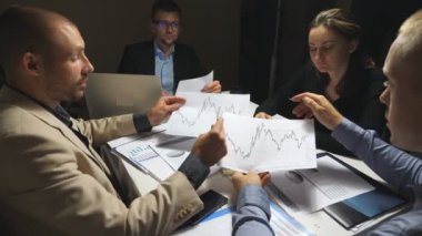 İş adamları iş günü sonunda kurumsal proje için strateji planlama. İş ekibi ofisteki masabaşında finansal grafikleri kontrol eder. Masada oturan ve belgeleri inceleyen genç iş arkadaşları