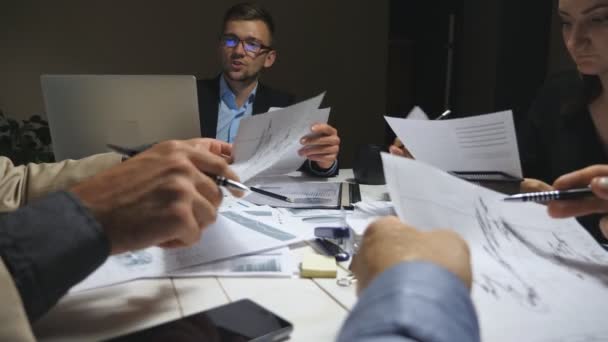 Jóvenes empresarios sentados a la mesa y discutiendo gráficos y gráficos de ingresos al final de la jornada laboral. Equipo de negocios examinando documentos en el escritorio. Colaboradores analizando informes financieros en la oficina — Vídeos de Stock