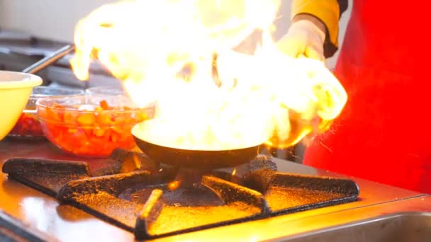 레스토랑의 현대적인 주방에서 팬에 남성 요리사 요리 플램브 스타일의 요리. 전문 요리사가 가스 플레이트에 불타는 프라이팬에 야채를 던지고 튀깁니다. 식사 준비 개념입니다. 슬로우 모션 — 비디오