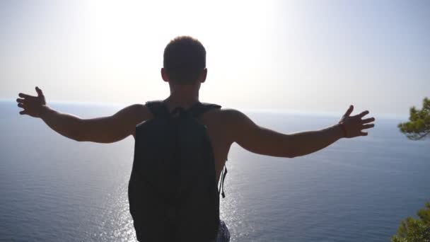 Caminante irreconocible con mochila de pie en el borde de la montaña y victoriosamente levantando las manos. Turista masculino descansando activamente durante el viaje disfrutando de la libertad y la hermosa vista del mar. Lento mo Vista trasera — Vídeo de stock