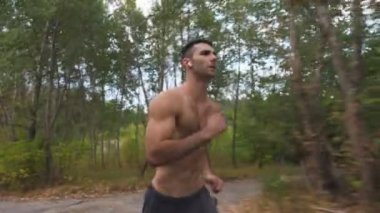 Kablosuz kulaklıkile yakışıklı kaslı adam erken sonbaharda orman yakınında iz boyunca hızlı sprint. Atlet adam doğada koşuyor. Genç sporcu antrenmanı açık havada. Sağlıklı aktif yaşam tarzı