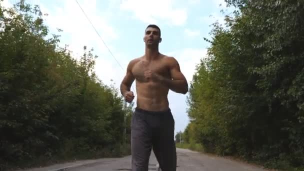 Guapo deportista corriendo rápido a lo largo de la carretera con un hermoso paisaje de fondo. Hombre musculoso joven con auriculares inalámbricos trotando a lo largo de la pista en la naturaleza. Vida activa saludable. Vista baja — Vídeos de Stock