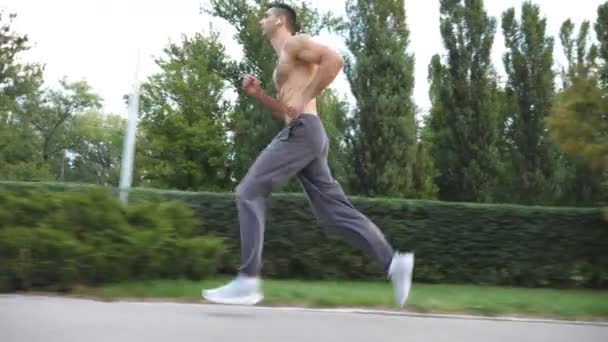 Joven deportista musculoso corriendo rápido a lo largo del sendero cerca del bosque en el día de verano. Atlético chico guapo con auriculares inalámbricos corriendo en la naturaleza. Concepto de estilo de vida activo saludable. Vista lateral Primer plano — Vídeos de Stock