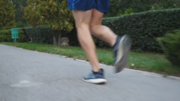 Homem bonito atlético com fones de ouvido sem fio correndo ao longo da calçada perto da floresta no dia de verão. Jovem desportista musculoso correndo na natureza. Conceito de estilo de vida ativo saudável. Guindaste tiro Close up — Vídeo de Stock