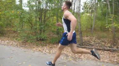 Kablosuz kulaklıklar ile yakışıklı güçlü sporcu orman yakınındaki iz boyunca hızlı sprint. Doğada yol boyunca koşan sportif bir adam. Atletik adam erken sonbaharda egzersiz. Sağlıklı aktif yaşam tarzı