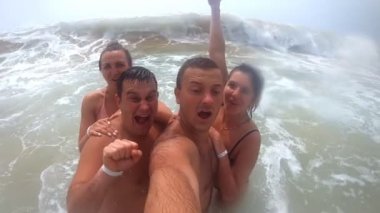 Deniz sahilinde kendi portresini çeken genç mutlu arkadaşlar. Büyük okyanus dalgası, kıyıda selfie çeken gülümseyen çiftleri kapsıyor. Neşeli erkek ve kadınlar tatil zevk ve deniz kenarında eğlenmek. Dinlenme kavramı