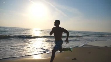 Gün doğumu sırasında sahil boyunca hızlı koşan genç sportif bir adamın silueti. Deniz sahilinde atletik çocuk eğitimi. Erkek sporcu açık havada egzersiz yapıyor. Sağlıklı aktif yaşam tarzı. Yan görünüm Yavaş hareket