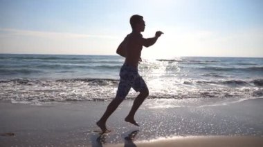 Deniz sahilde sabah koşan genç sportif adam. Sahil boyunca güneşli bir günde koşu atletik bir adam. Açık egzersiz erkek sporcu. Sağlıklı aktif yaşam tarzı. Yan görünümü ağır çekim