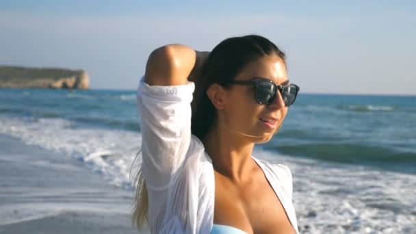 Hermosa chica en gafas de sol sonriendo y jugando con su pelo rubio en la costa. Mujer bronceada disfrutando de la vida y divirtiéndose en la orilla. Concepto de vacaciones de verano o vacaciones. Movimiento lento — Vídeos de Stock