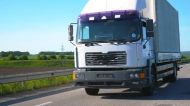 Kamera, kargo karavanıyla otoyolda giden ve yaz günü malları taşıyan kamyonu takip ediyor. Arka planda doğa manzarası olan bir kamyon. Yavaş çekim Ön görünüm Kapat