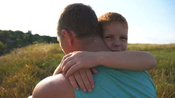 Pequeño chico pelirrojo sentado en la hierba verde en el césped con su padre y abrazándolo con amor. Feliz hijo abrazando a su padre en el prado en un día soleado. Familia pasar tiempo juntos en la naturaleza . — Vídeos de Stock