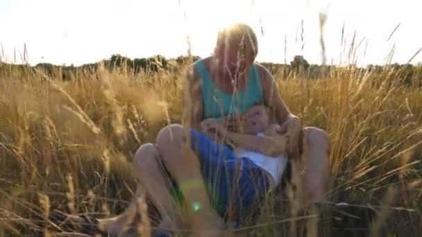 Счастливый отец и его сын сидят на высокой траве на поле и веселятся вместе в солнечный день. Веселый отец, держащийся за шип и играющий со своим ребенком на лугу. Закрыть Медленное движение — стоковое видео