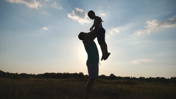 Šťastný tatínek hází svého chlapečka do vzduchu v přírodě. Siluety otce a syna hrají na louku při západu slunce. Otec a dítě tráví čas pohromadě a baví se venku. Zpomaleně — Stock video