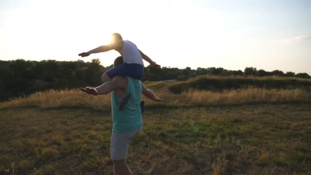 Kleine jongen zittend op de schouders van zijn vader en het spelen van verhoogde handen als vliegtuig buiten. Vader draagt zoon en verhoogde armen als vliegtuig op weide. Familie tijd samen doorbrengen in de natuur. Achteraanzicht — Stockvideo