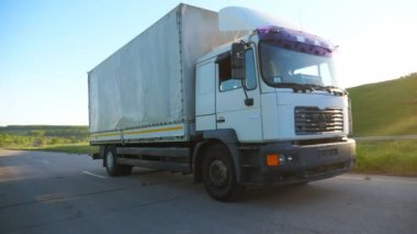 Yük römorklu kamyon karayolu üzerinde sürme ve gün batımında mal taşıma. Güzel doğa ortamı arasında kırsal yol üzerinden kamyon sürme. Arka planda parlayan parlak güneş. Yavaş çekim