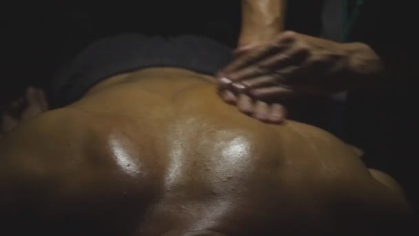 Esportista muscular deitado na mesa de massagem e braços de massagista massageando os ombros em um quarto escuro. Mãos masculinas de massagista profissional fazendo rubdown cura de volta ao homem com óleo no salão — Vídeo de Stock