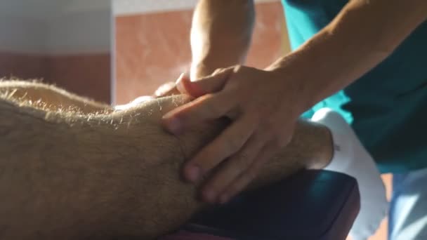 Le braccia vicine di massaggiatore professionale che fanno lentamente sfregano la gamba di atleta che si trova su tavolo di massaggio in salone. Mani maschili di massaggiatore guarigione massaggiando piede di giovane sportivo in salotto. Rallentatore — Video Stock