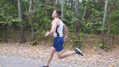 Kablosuz kulaklıklar ile yakışıklı güçlü sporcu orman yakınındaki iz boyunca hızlı sprint. Doğada yol boyunca koşan sportif bir adam. Atletik adam erken sonbaharda egzersiz. Sağlıklı aktif yaşam tarzı