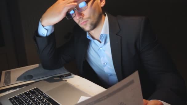 Joven hombre de negocios cansado sentado en la mesa y analizando el informe financiero en el cargo. Empresario molesto trabajando en la computadora portátil tarde en la noche posponiendo las gafas y frotándose los ojos. Movimiento lento — Vídeos de Stock