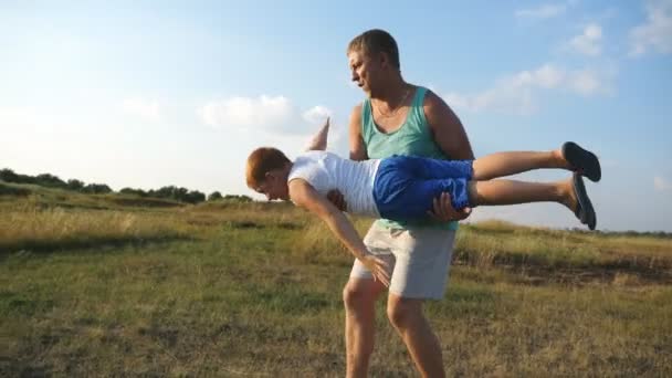 Père joyeux tenant et tournant son petit enfant dans les mains. Le petit roux imagine qu'il vole comme un avion. Heureux père et fils jouant sur la prairie à la journée ensoleillée. La famille passe du temps dehors. Moteur lent — Video