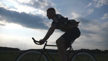 Arka planda güzel günbatımı gökyüzü ile vintage bisiklet sürme genç adam silueti. Kırsal yolda sportif adam bisiklete biner. Erkek bisikletçi sabit vitesli bisiklet sürme. Aktif yaşam tarzı Yavaş hareket