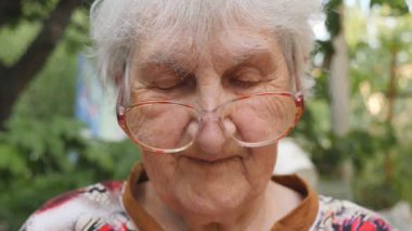 Gözlüklü yaşlı kadın kameraya bakıyor ve dışarıda gülümsüyor. Gözlüklü mutlu büyükannenin portresi dışarıda vakit geçiriyor. Büyükannenin duyguları. Yavaş hareketi kapat