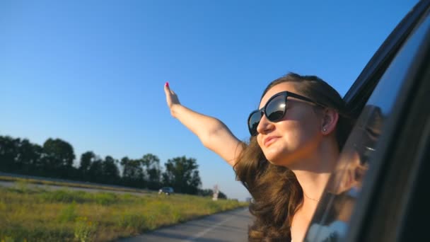 Gelukkig meisje in zonnebril leunend uit auto raam en genieten van de reis. Jonge vrouw kijkt uit raam van bewegende auto op zonnige dag. Reizen en vrijheid concept. Slow motion close-up — Stockvideo